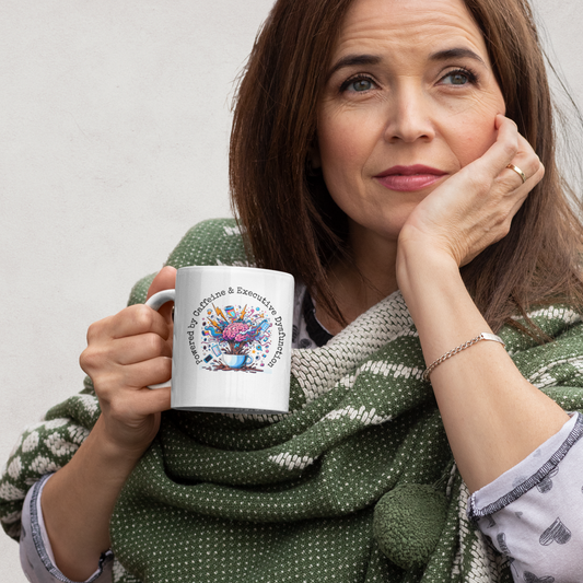 Woman wrapped in a cozy sweater holding a mug with the quote “Powered by Caffeine & Executive Dysfunction” – Funny ADHD mug by Mentally Glitched.